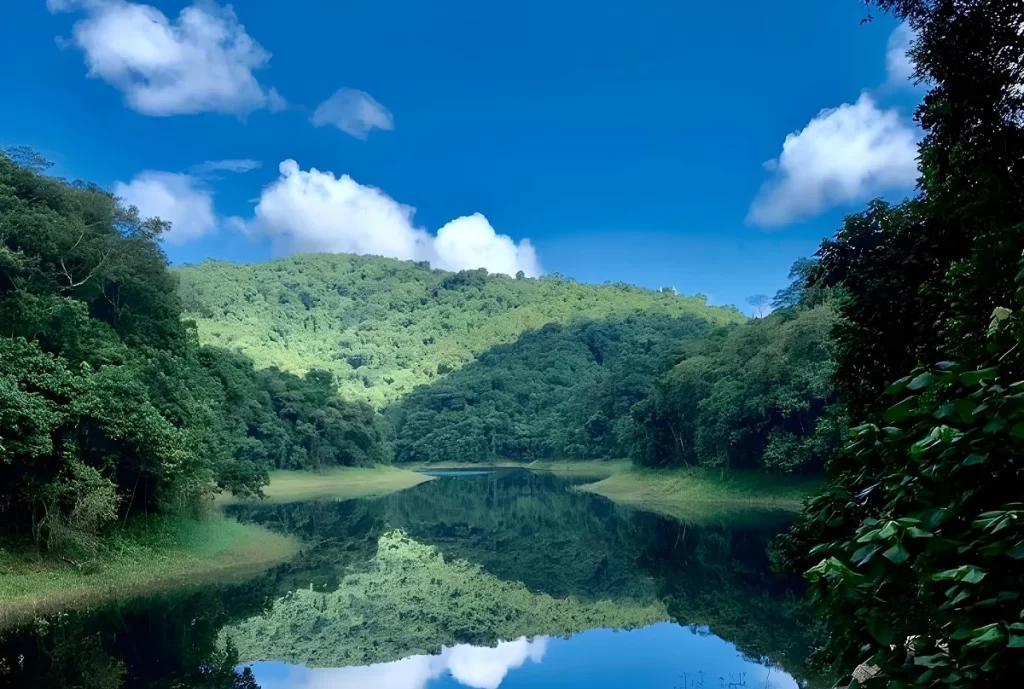 Paisagem exuberante da Floresta da Cantareira em Mairiporã, mostrando o reflexo das montanhas verdes e do céu azul com nuvens brancas nas águas calmas da Represa Paulo de Paiva Castro.