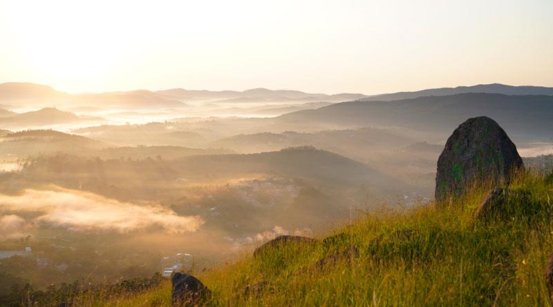 Fotografia panorâmica do Pico do Olho D’água em Mairiporã, exibindo o nascer do sol sobre as colinas cobertas de neblina e um gramado verde em primeiro plano, com uma grande rocha à direita.